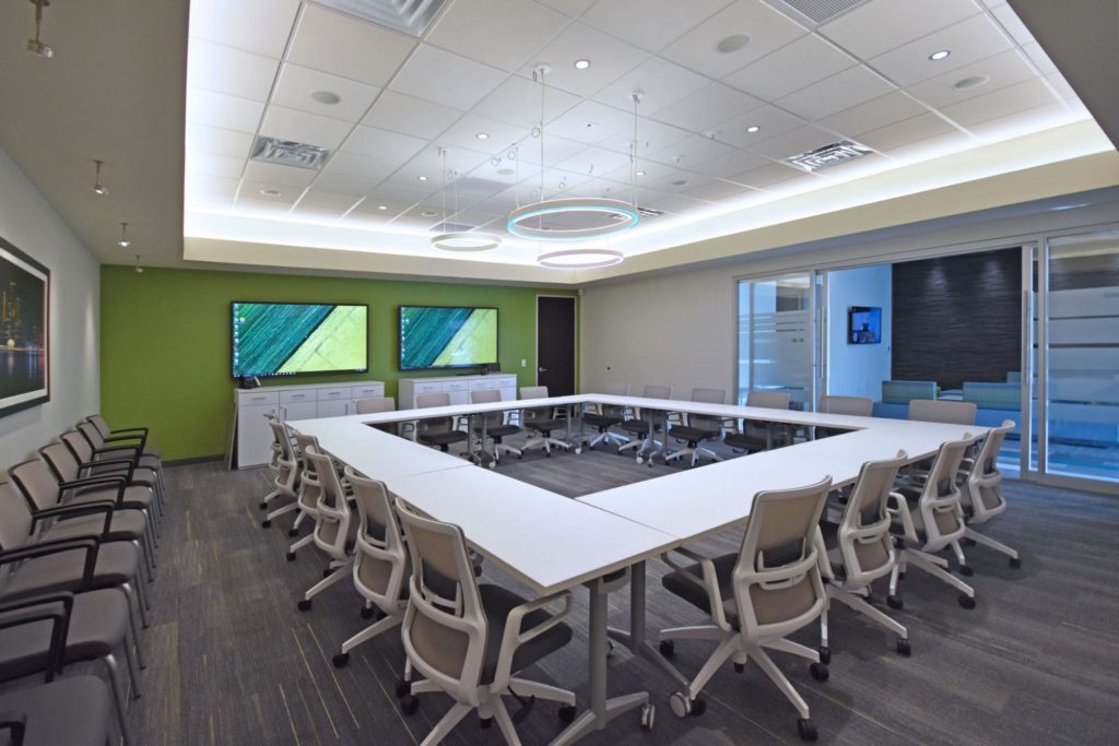 Office conference room with tables arranged in a square shape next to two video monitors on the wall.