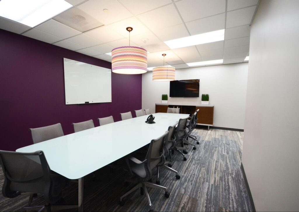 Conference room with long table, purple accent wall, whiteboard and video monitor.